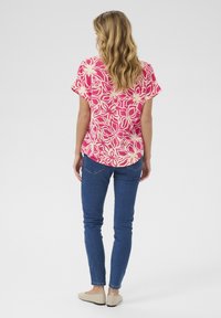 Top rosa a maniche corte con motivo floreale, abbinato a jeans skinny blu e scarpe beige slip-on. Vista posteriore, visibili lunghi capelli mossi.