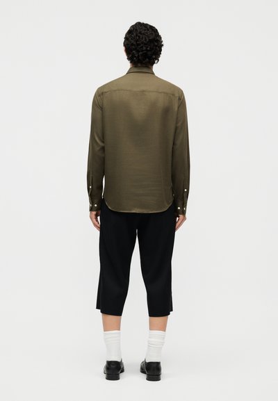 Άτομο που φαίνεται από πίσω, φορώντας πουκάμισο μακρυμάνικο σε ελαιδί πράσινο, μαύρο παντελόνι μέχρι τον αστράγαλο, λευκές κάλτσες και μαύρα παπούτσια, μπροστά σε απλό φόντο.