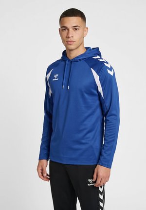 Młody mężczyzna ubrany w niebieską sportową bluzę z kapturem marki hummel z białymi pasami w kształcie jodełki oraz czarne spodnie hummel z białymi pasami w kształcie jodełki po bokach.