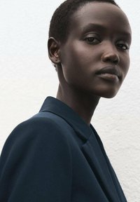 Marineblauwe blazer met gestructureerde revers en gladde stof, gedragen door een model met kort, nauwkeurig geknipt haar tegen een gestructureerde witte achtergrond.