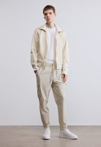 Ελαφρύ μπεζ μπουφάν με φερμουάρ και υφή, λευκό μπλουζάκι και ελαφριά μπεζ παντελόνια cargo. Διαθέτει ελαστική μέση και πλαϊνές τσέπες.