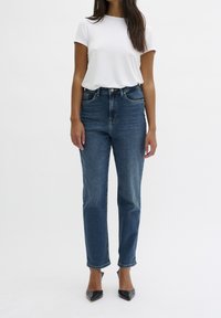 Vaqueros de cintura alta y pierna recta en denim azul oscuro, con cinco bolsillos, trabillas para el cinturón y ligero desgaste en el dobladillo. Combinados con una camiseta blanca.