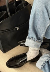 Black leather loafers with shiny finish, gray ribbed socks, and light blue cuffed denim jeans. A black leather tote bag is in the background.