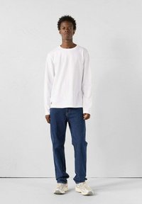 Camicia bianca a maniche lunghe abbinata a jeans blu e sneakers di colore chiaro. Design semplice con una vestibilità rilassata e senza motivi o accenti visibili.