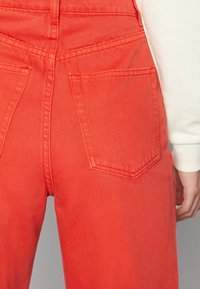 Rote Denim-Latzhose mit glatter Textur, die zwei aufgesetzte Taschen auf der Rückseite und sichtbare Nähedetails aufweist.