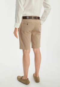 Beżowe spodenki cargo o prostym kroju, z dwiema tylnymi kieszeniami i przeszyciami. Noszone z jasnym koszulą i brązowym paskiem.