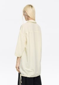 Camicia oversize gialla chiara con colletto, maniche corte e orlo arricciato. Il tessuto sembra morbido e leggermente strutturato.