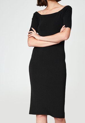 Femme portant une robe noire ajustée mi-longue avec des manches courtes et un large décolleté carré, bras croisés devant.
