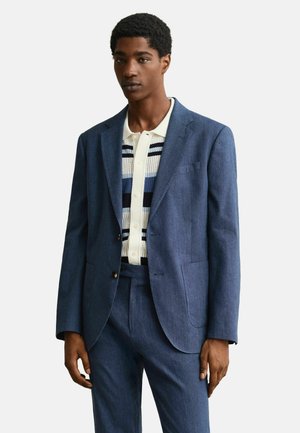 Man in een blauw pak met bijpassende broek en een witte gebreide polo met blauwe en zwarte strepen, staand tegen een effen achtergrond.