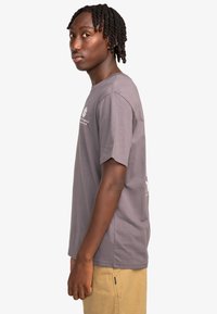 Jongen met gevlochten haar, gekleed in een grijze T-shirt met korte mouwen en beige shorts, staand in profiel tegen een witte achtergrond.