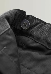 Nærbillede af en sort knap syet fast på en stofløkke, som er fastgjort til et sort og mørkegråt tekstureret stof.