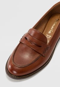 Mocassin en cuir marron avec un bout arrondi, une couture décorative et une bride surélevée traversant le cou-de-pied. Intérieur texturé et semelle contrastée.