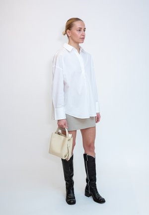 Nainen, jolla on tukka nutturalla, pukeutuu ylisuurioon valkoiseen paitaan, beigeen minihameeseen, mustiin polven yli ulottuviin saappaisiin ja hän pitää beigeä käsilaukkua yksiväristä taustaa vasten.