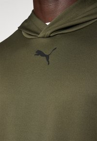 Maslinasto zeleni dukserica od mekanog materijala, sa kenguruh džepom i crnim logotipom skakačkog pumasa na prednjem dijelu.