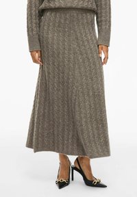 Lange, ribbelige lichtbruine gebreide rok met een uitlopende zoom, gecombineerd met zwarte slingback hakken met spitse neuzen en een gouden kettingdetail.