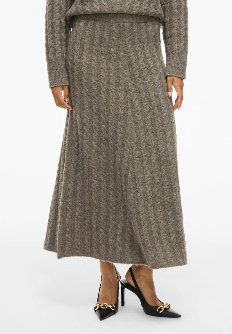 Lange, ribbelige lichtbruine gebreide rok met een uitlopende zoom, gecombineerd met zwarte slingback hakken met spitse neuzen en een gouden kettingdetail.