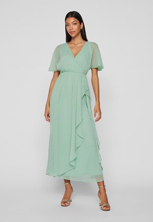 Woman wearing a floor-length, light green wrap dress with flutter sleeves, ruffle details, and gold high-heeled sandals, standing against a gray background.