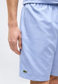 Δεξί χέρι δίπλα σε ανοιχτό μπλε σορτς με κεντημένο πράσινο λογότυπο κροκόδειλου κοντά στο τελείωμα σε απλό φόντο.