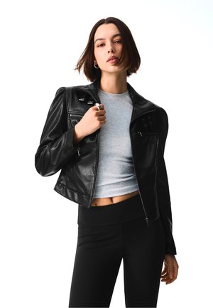 Jeune femme aux cheveux courts et bruns porte une veste en cuir noire sur un crop top gris et un pantalon noir, tenant le revers de la veste.