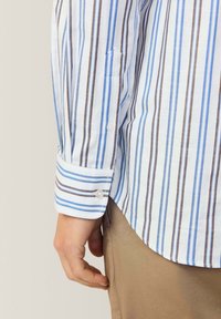 Camicia di cotone con righe verticali blu e marroni, dotata di polsino con bottone e taschino sul petto. Vestibilità sagomata, orlo arrotondato.