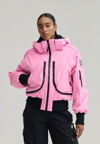 Nő hátrafogott hajjal, rózsaszín cipzáras dzsekit visel fekete betétekkel és fekete nadrágot, sima háttér előtt áll.