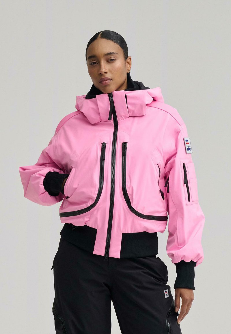 Nő hátrafogott hajjal, rózsaszín cipzáras dzsekit visel fekete betétekkel és fekete nadrágot, sima háttér előtt áll.