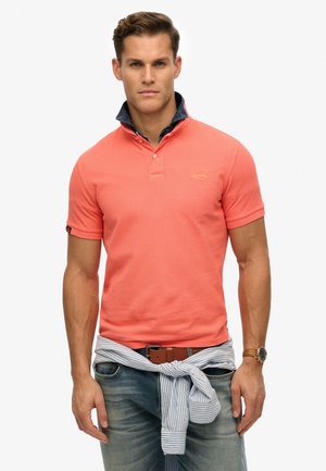 Camisa polo de color coral con cuello abotonado y un interior de cuello de mezclilla. Combinada con unos vaqueros de color azul claro y una tela a rayas atada alrededor de la cintura.