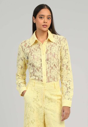 Femme portant une chemise boutonnée à manches longues en dentelle jaune clair et un pantalon taille haute assorti en dentelle, debout devant un fond uni clair.