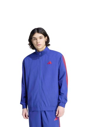 Un mannequin porte un survêtement bleu orné de bandes et de logos Adidas rouges sur fond blanc.