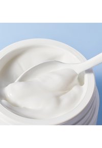 Vit kräm i en rund, vit burk med slät textur, delvis utgott med en vit sked mot en ljusblå bakgrund.