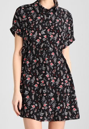 Femme portant une robe noire à manches courtes ornée de petits motifs floraux rouges, blancs et gris, boutonnée à l'avant, debout devant un fond uni.