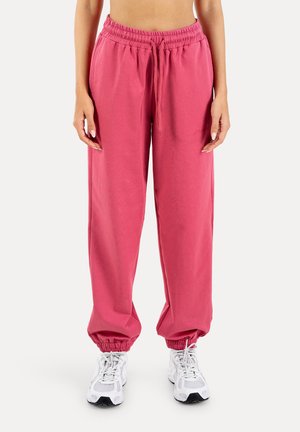 Donna che indossa pantaloni jogger con elastico in vita rosa e scarpe da ginnastica bianche, in piedi davanti a uno sfondo chiaro e uniforme.