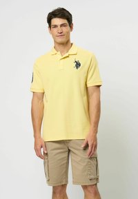 Žltá polo košela vyrobená z texturovanej bavlny, s námořníckym logom na ľavej hrudi a krátkymi rukávmi, spojená s béžovými cargo šortkami.