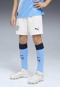 Equipación de fútbol para niños del Manchester City: camiseta celeste con detalles en blanco, pantalones cortos blancos con logo negro de Puma, medias azules con banda oscura y logo, botas plateadas.