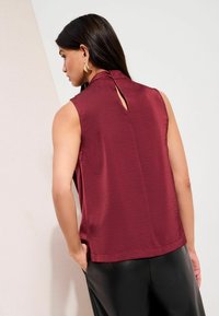 Bordeauxrote ärmellose Bluse mit hohem Kragen und Schlitzdetail am Rücken, aus einem strukturierten Stoff, kombiniert mit schwarzen Hosen.