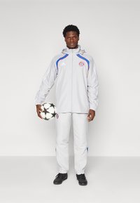 Grijze sportjack met rode en blauwe accenten, bijpassende grijze broek, houdend een zwart-wit gestreepte voetbal, zwarte schoenen.