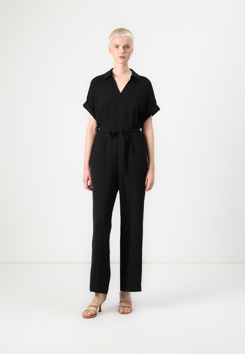 Svart jumpsuit med V-hals, korte oppbrettete ermer, belte i livet og brede rette ben. Brukt med beige hæl sandaler.