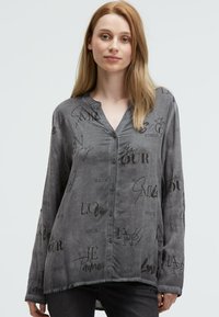 Junge Frau mit langen blonden Haaren, die ein graues Hemd mit schwarz geschriebener Schrift und dunklen Hosen vor einem hellen Hintergrund trägt.