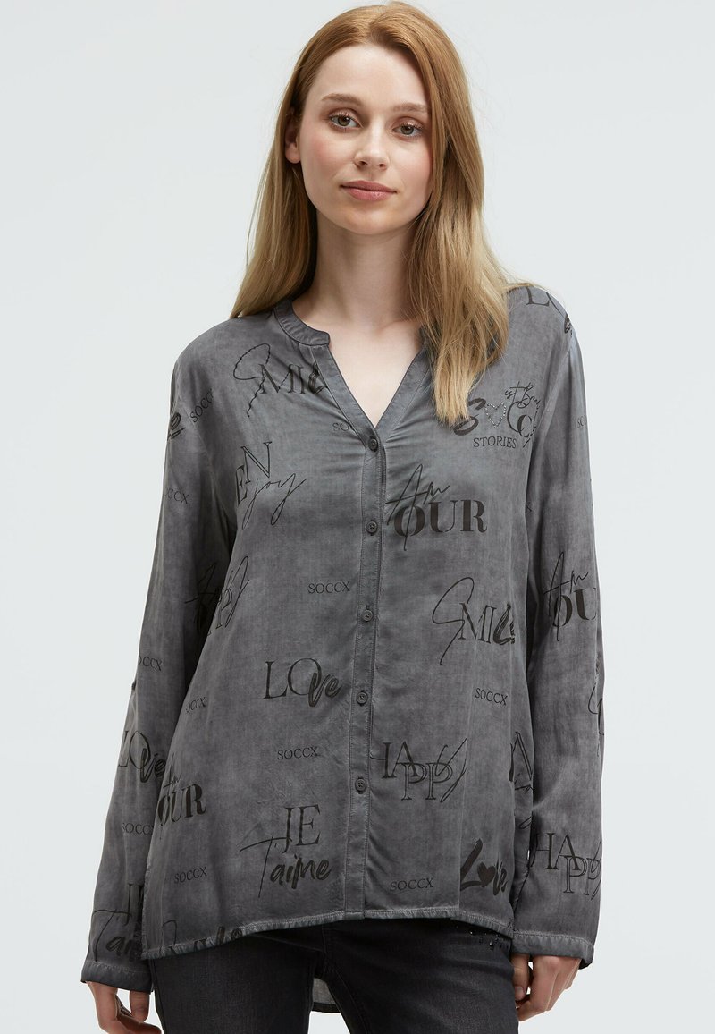 Junge Frau mit langen blonden Haaren, die ein graues Hemd mit schwarz geschriebener Schrift und dunklen Hosen vor einem hellen Hintergrund trägt.