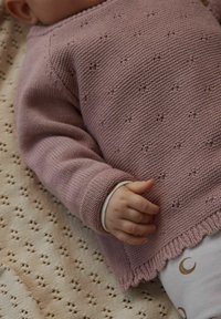 Roza pleten pulover z reliefnim vzorcem, s podaljšanimi manšetami in valovitim robom, nošen čez svetla oblačila z vzorci polmeseca.