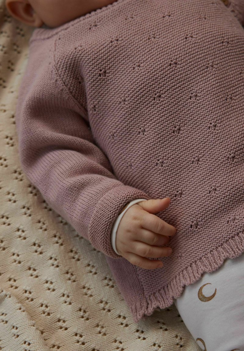 Roza pleten pulover z reliefnim vzorcem, s podaljšanimi manšetami in valovitim robom, nošen čez svetla oblačila z vzorci polmeseca.