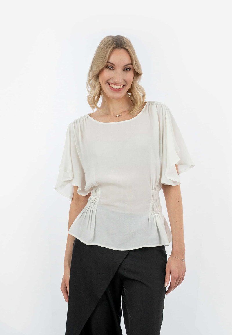 Blusa bianca con lati arricciati, maniche fluttuanti corte e vestibilità rilassata. Abbinata a pantaloni neri a gamba larga.