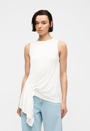 Femme aux cheveux courts et foncés portant un haut blanc sans manches avec un détail drapé froncé et un pantalon bleu clair à fines rayures, debout devant un fond uni.