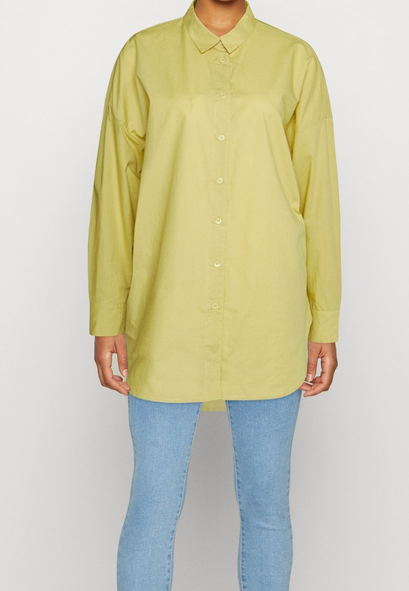 Camicia a maniche lunghe in cotone giallo chiaro. Presenta un colletto, bottoni frontali e una vestibilità rilassata. Indossata con jeans blu chiaro.