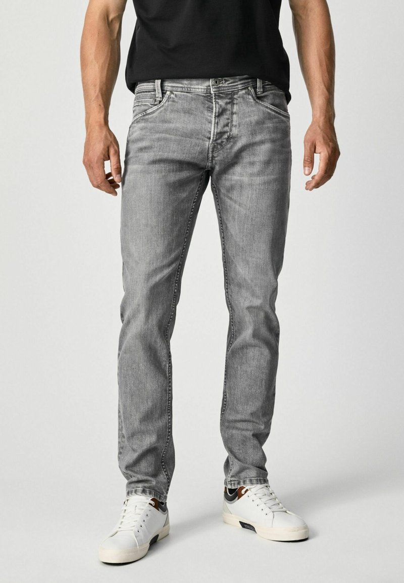 Hombre que lleva puestos unos jeans ajustados de color gris claro y zapatillas blancas, de pie frente a un fondo liso claro.
