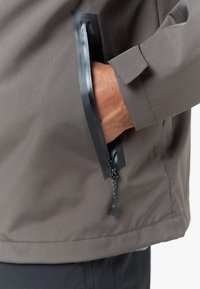 Vodootporna siva jakna s patentnim zatvaračem, koja ima džep s crnim akcentima, glatku teksturu i elegantan, moderni dizajn.