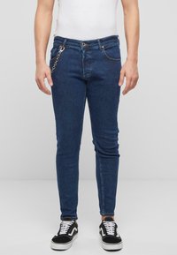 Slim-fit siniset farkut, jotka on valmistettu denimistä. Niissä on vetoketjulla ja napilla suljettava etuosa, viisi taskua ja metalliketjuverhoilu.