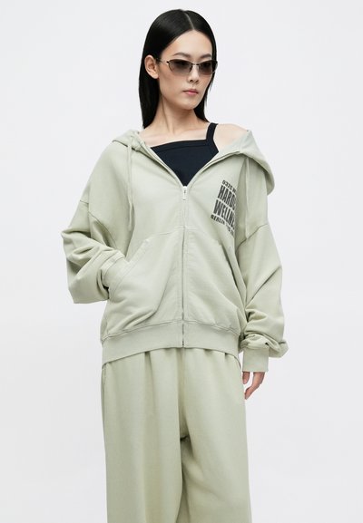 Jasnozielona bluza z kapturem i zamkiem, w oversize'owym kroju, z kieszenią z przodu i nadrukowanym tekstem. Nosi na czarnym topie i dopasowanych luźnych spodniach.