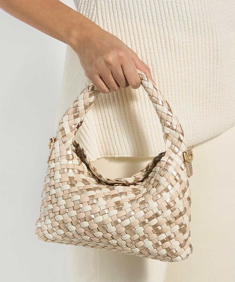 Main tenant un sac à main tissé beige et blanc avec une anse tressée, devant un haut crème texturé et un pantalon clair.