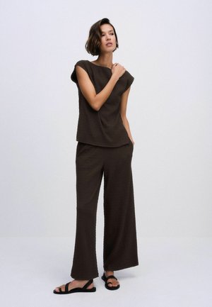 Kobieta w ciemnobrązowym topie bez rękawów i szerokich spodniach z czarnymi sandałami, stojąca na jednolitym jasnoszarym tle.